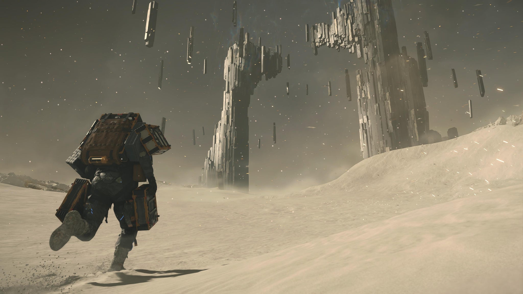 Death Stranding 2 screenshot showing Sam carrying backpack into a desert environment with strange monoliths in the background