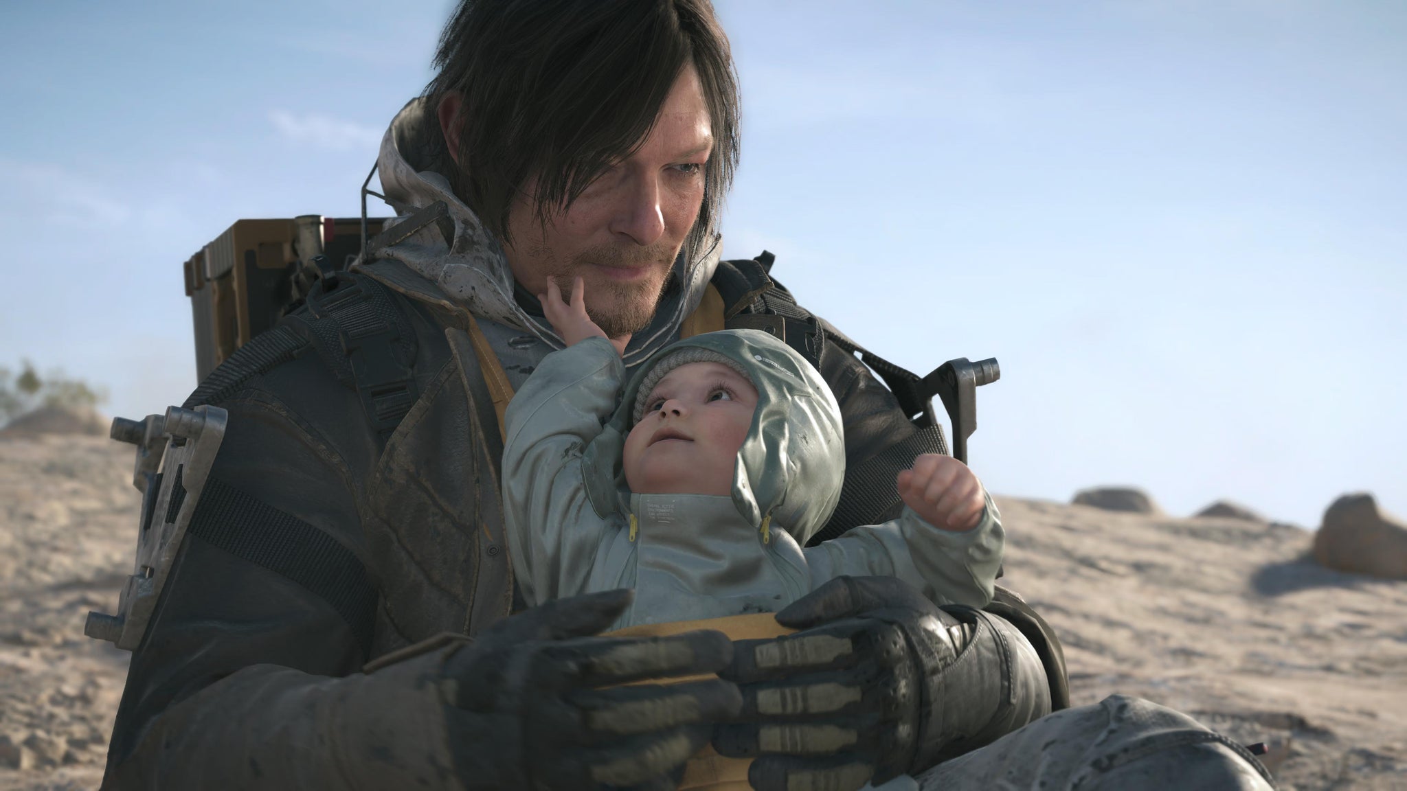 Death Stranding 2 screenshot showing close up of Sam holding a baby while sat in a desert